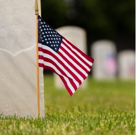 A small American flag on a stick stands in front of a military cemetery headstone | BRG