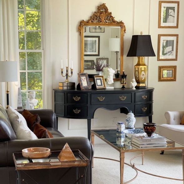 Ebonized server table with brass pulls in Nancy's living room. Displayed are framed photos, sculptures, candelabra, lamp, and mirror. Gilt artwork and furniture complete the elegant space. | BRG