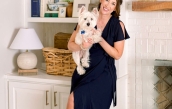 Image of Sally and her puppy smiling in front of a built-in shelf and fireplace, adorned with decorative items such as table lamps, candlesticks, blue and white porcelain pieces, photo frames, and books, against a backdrop of white-painted walls.  | BRG